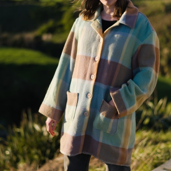 Recycled wool blanket coat A photograph of a woman wearing a wool coat. It goes to her knees and features pockets and large cream coloured buttons. The coat is made of a rainbow coloured wool blanket fabric.