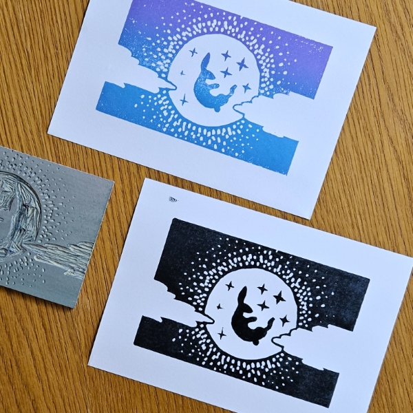 Linocut design & prints Two printed linocut designs next to the original linocut. The linocut design is a rabbit in front of the moon with stars, shining against a dark sky with clouds slightly obscuring either side. One print is black, and one is ombre purple and blue.
