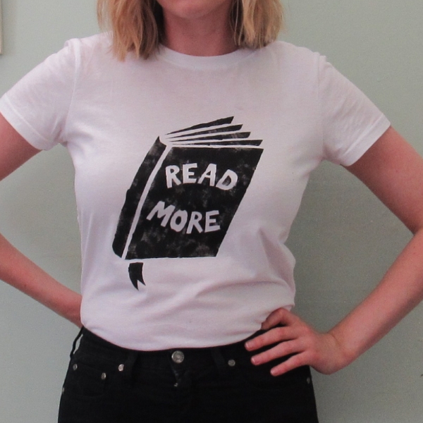 Read More t-shirt design A close up photograph of the torso of a woman, showing the t-shirt design. The design features a black screen printed book with the words 'Read More' as the title text, on a white t-shirt.